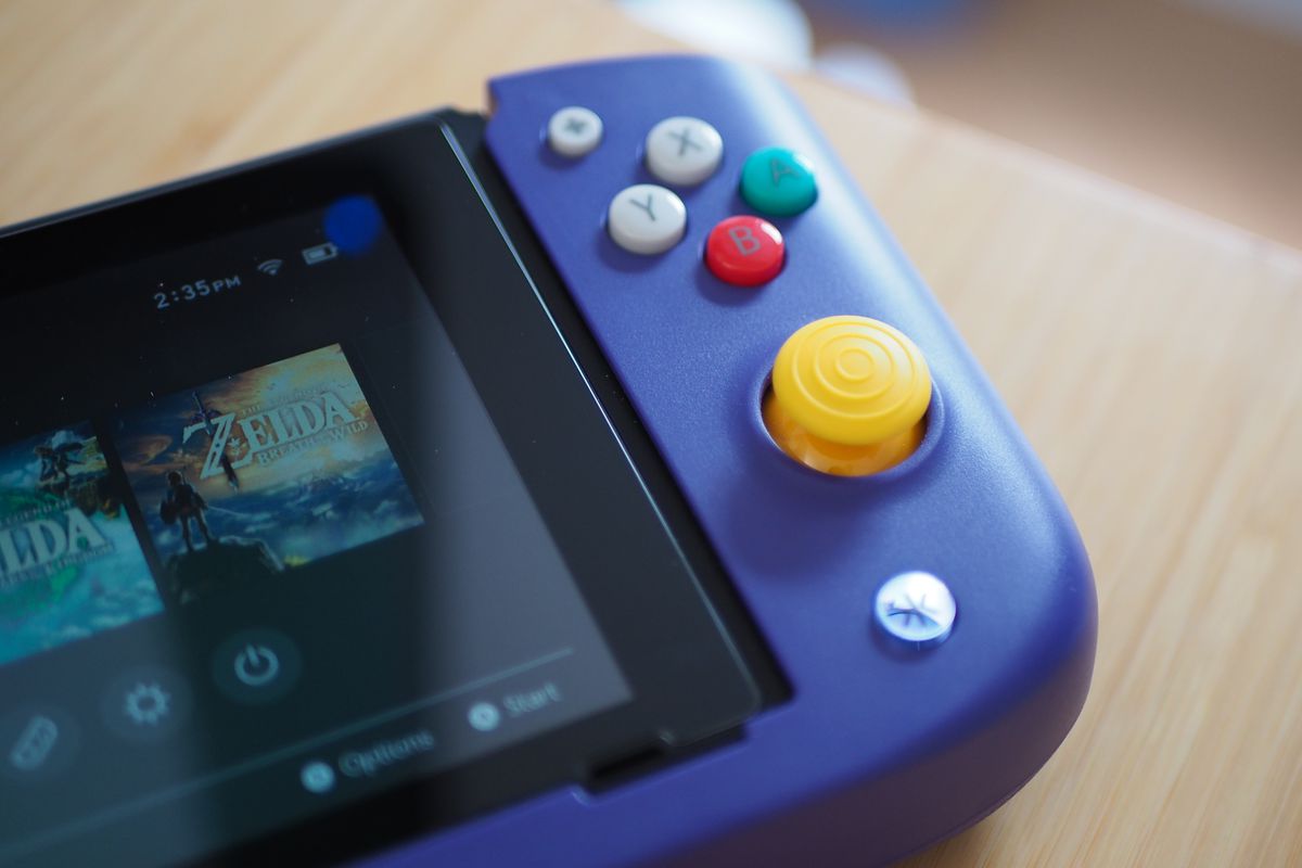 Crkd Nitro Deck کے دائیں جانب کی ایک تصویر، ایک Nintendo Switch ہولڈر جس میں کنسول کے تمام کنٹرولز اور پورٹس ہیں۔ یہ نائنٹینڈو گیم کیوب کی نقل کرتے ہوئے رنگین جوائس اسٹک اور بٹنوں کے ساتھ جامنی ہے۔