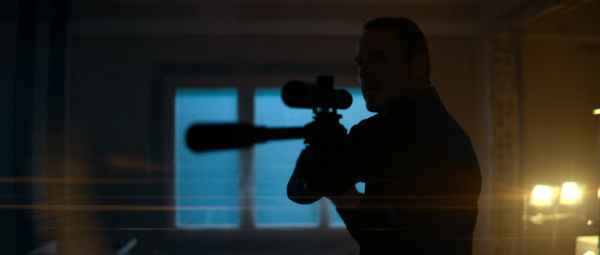Michael Fassbender as an assassin pointing a sniper rifle through a window in The Killer.