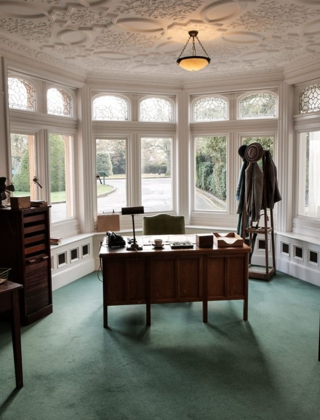 A photo of Alastair Denniston's office at Bletchley Park, containing an old-fashioned wooden desk, chair, lamp, and coat rack