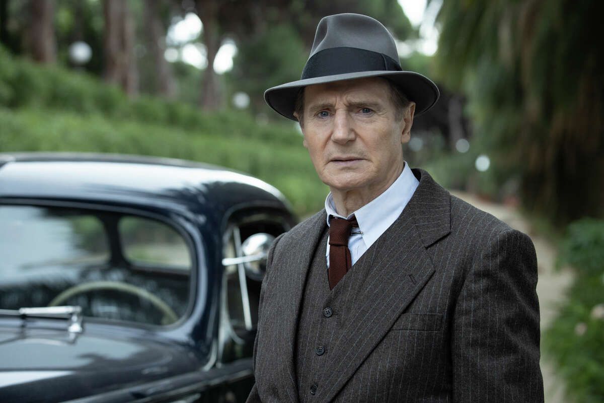 Mężczyzna (Liam Neeson) stojący w zalesionej okolicy przed ciemną sukienką typu sedan w brązowym prążkowanym garniturze, ciemnoczerwonym krawacie i szarej fedorze.