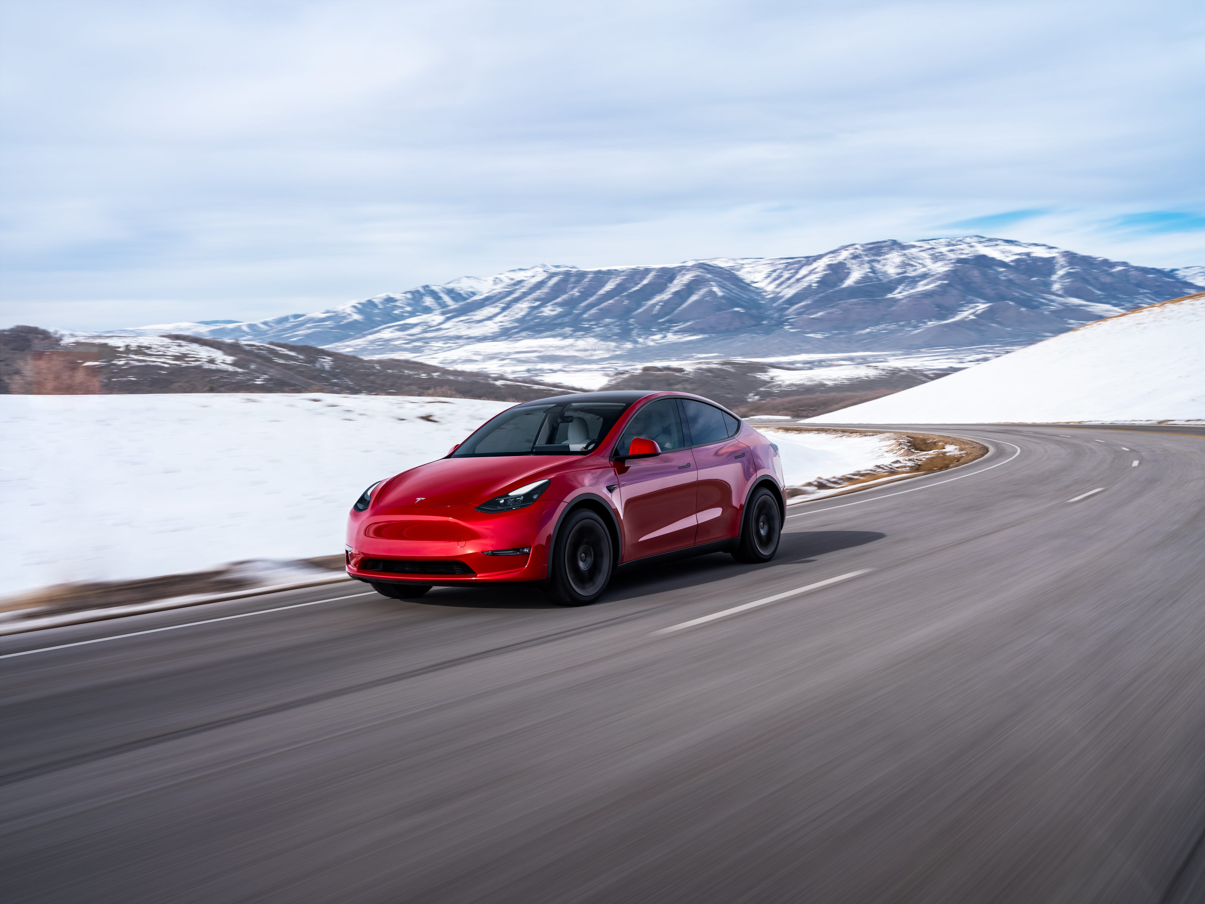 Um SUV Tesla Model Y vermelho dirige por uma estrada com neve e montanhas ao fundo.