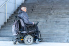 An electric wheelchair user looks up at an outdoor staircase. No ramp or other alternative access is visible