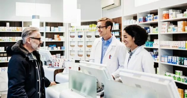 Two pharmacists stood behind the counter, demonstrating excellent customer service, talking to a regular customer who has come to pick up his prescription