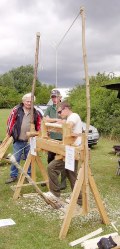A bodger's pole lathe at an event. The piece being turned is in a wooden frame, either side of which is a springy branch. A rope goes between the tops of the branches, and another passes from its centre downwards and round the piece to be worked, to a treadle which the operator moves with his foot to operate the lathe.