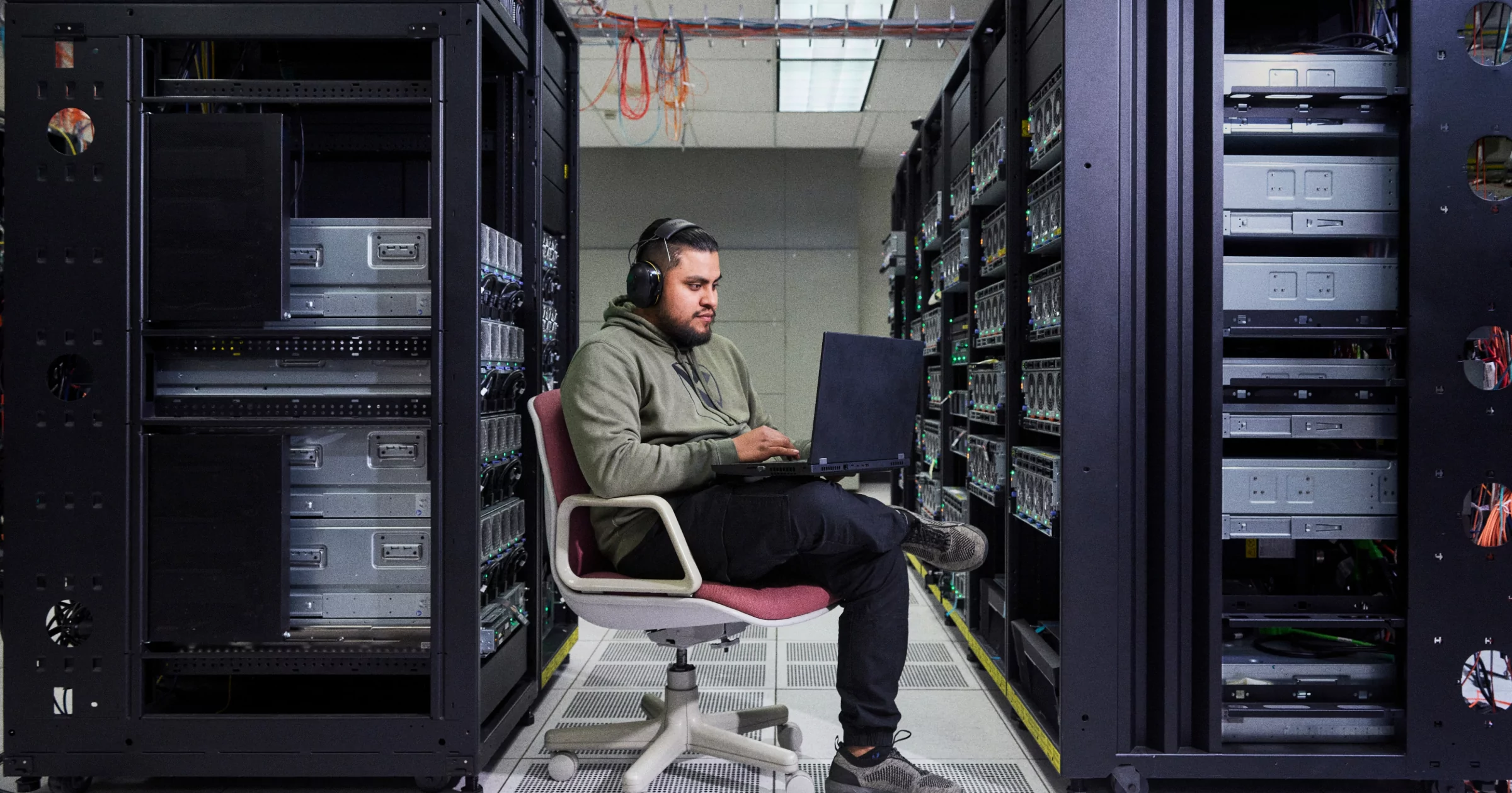 Developer sitting in chair in between storage shelves working on laptop and wearing headphones and smiling