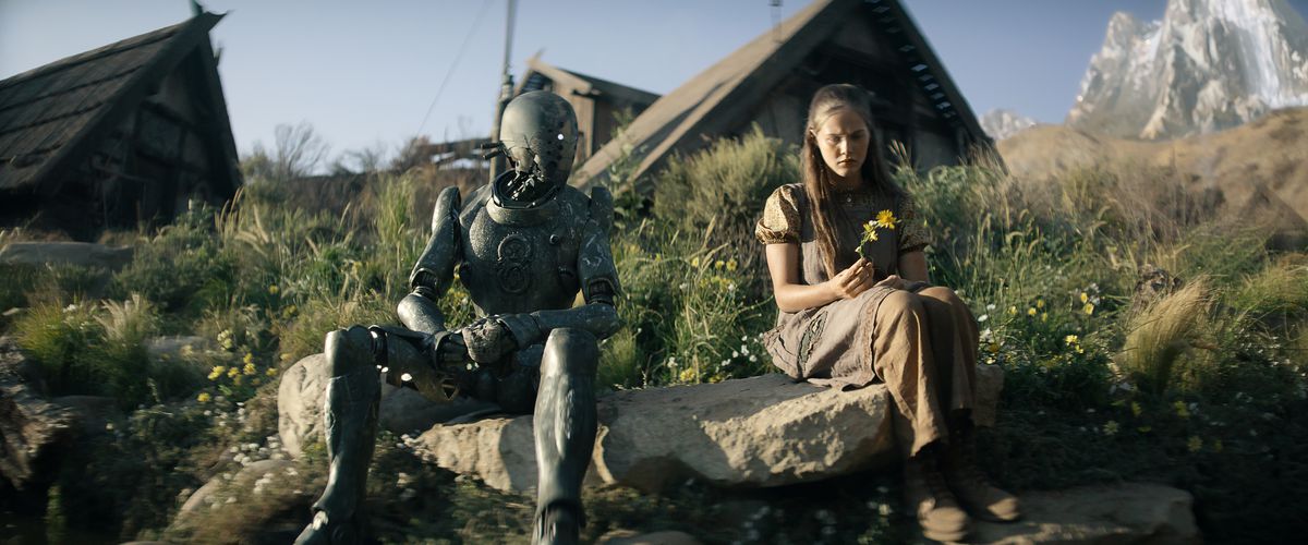 Un robot humanoïde est assis sur une colline verdoyante devant des tentes, à côté d'une jeune femme à l'air triste qui tient une fleur jaune dans Rebel Moon Part One: A Child of Fire.