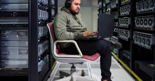 Henkilö istuu tuolilla tietovarastossa kuulokkeet päällään ja työskentelee kannettavalla tietokoneella