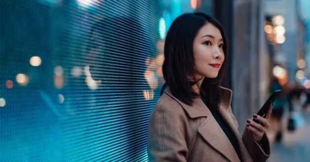 Ung asiatisk forretningskvinde bruger mobiltelefon, mens hun venter på bygaden om natten. Mod et massivt digitalt display med bylys oplyst i baggrunden. Forbind fremtiden.
