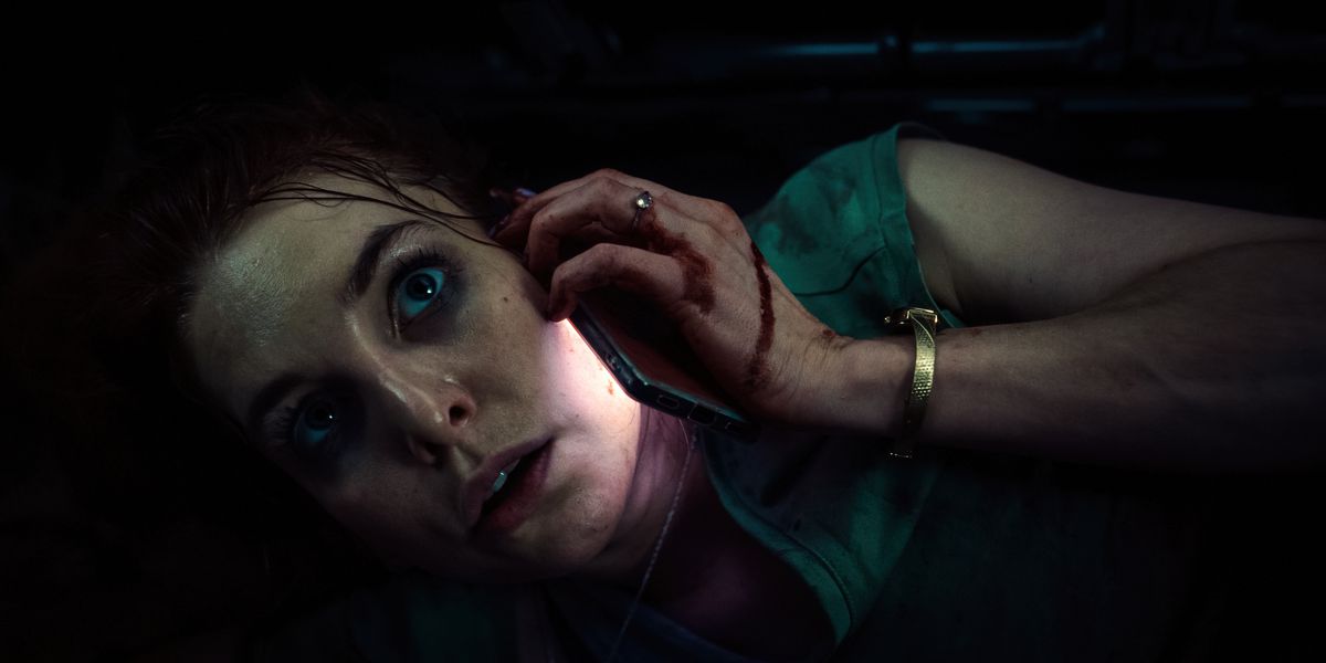 Uma mulher com sangue seco na mão segura um telefone no rosto enquanto está presa no porta-malas de um veículo.