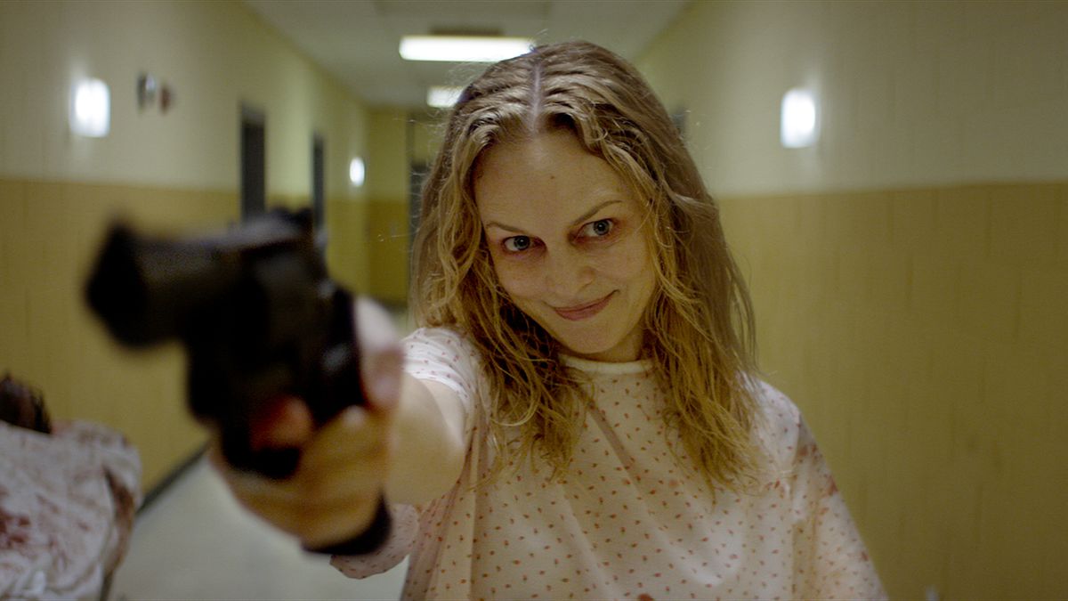 Una donna sorridente in camice da ospedale punta una pistola in un corridoio scarsamente illuminato in Carne adatta.