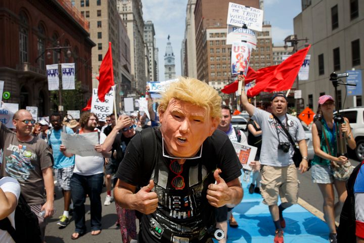 Un bărbat poartă o mască de față Donald Trump în timp ce oamenii marșează ținând pancarte în sprijinul lui Bernie Sanders în timpul unui protest la convenția democrată.