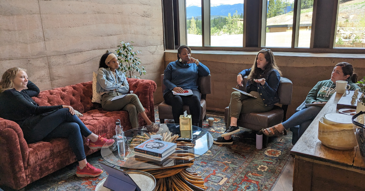 Five women sit around a coffee table with mountains behind them watching one of the women speak