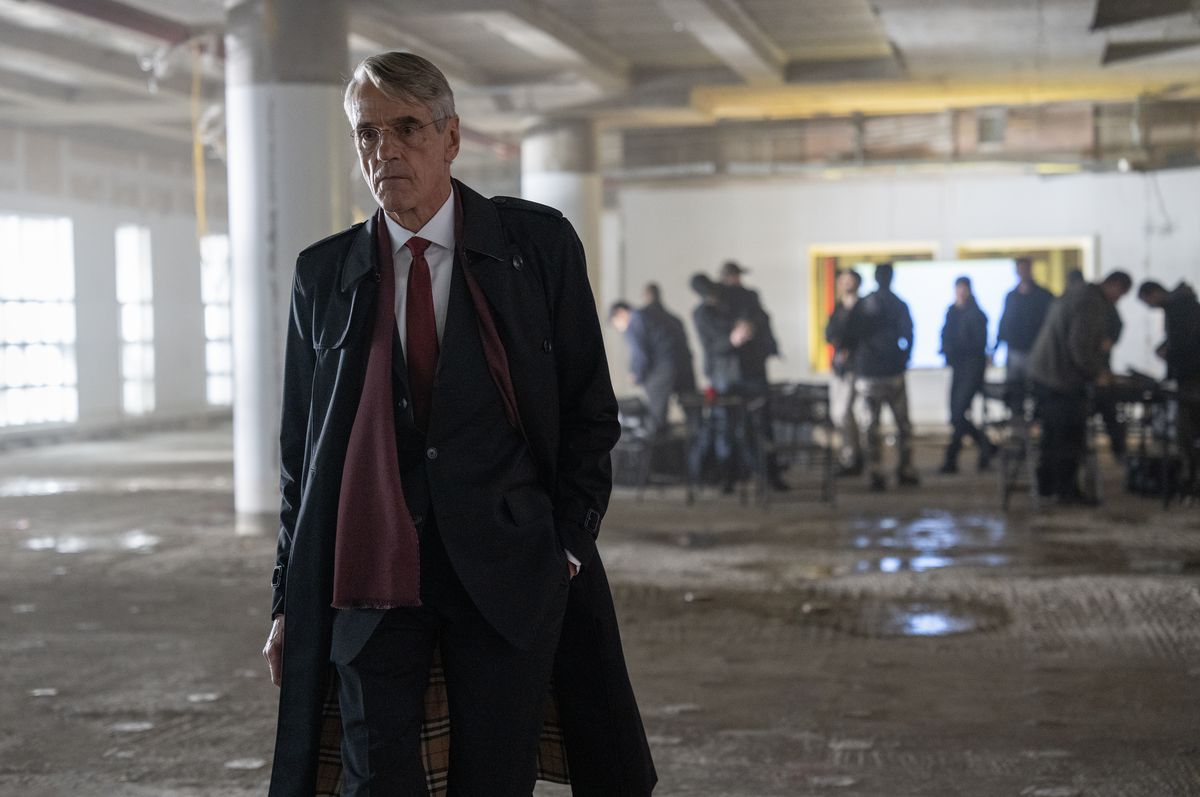 Jeremy Irons, wearing a long coat, walks towards the camera and away from uniformed people in what looks like a parking garage in The Beekeeper.