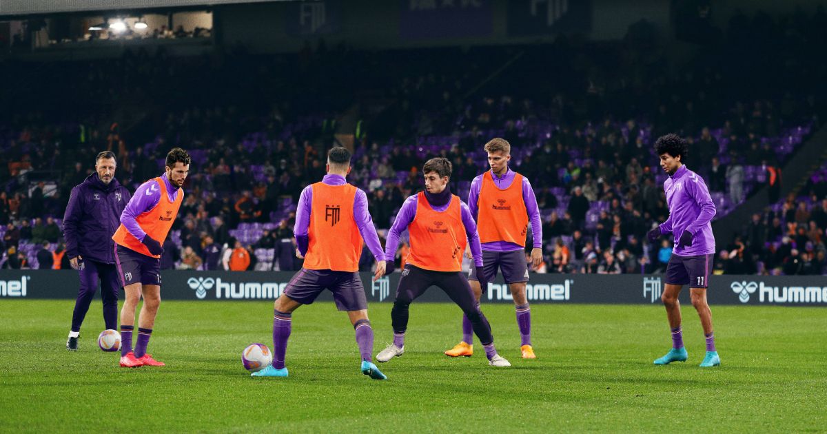 A team in purple training kit and orange bibs practicing football in front of a manager in a stadium.