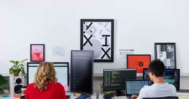 Dua pengembang duduk di kursi meja menghadap dinding mengerjakan komputer
