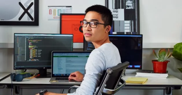 Ung person med briller sitter i skrivebordsstol foran tre dataskjermer, vendt mot kameraet og smiler
