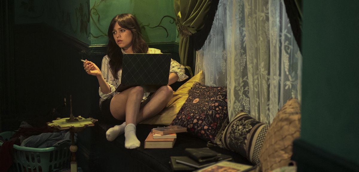 Cairo (Jenna Ortega) ngồi hút thuốc với chiếc máy tính xách tay trên đùi và hai chân xếp thành ghế, xung quanh là gối, sách và một tấm rèm ren mỏng trong Miller's Girl