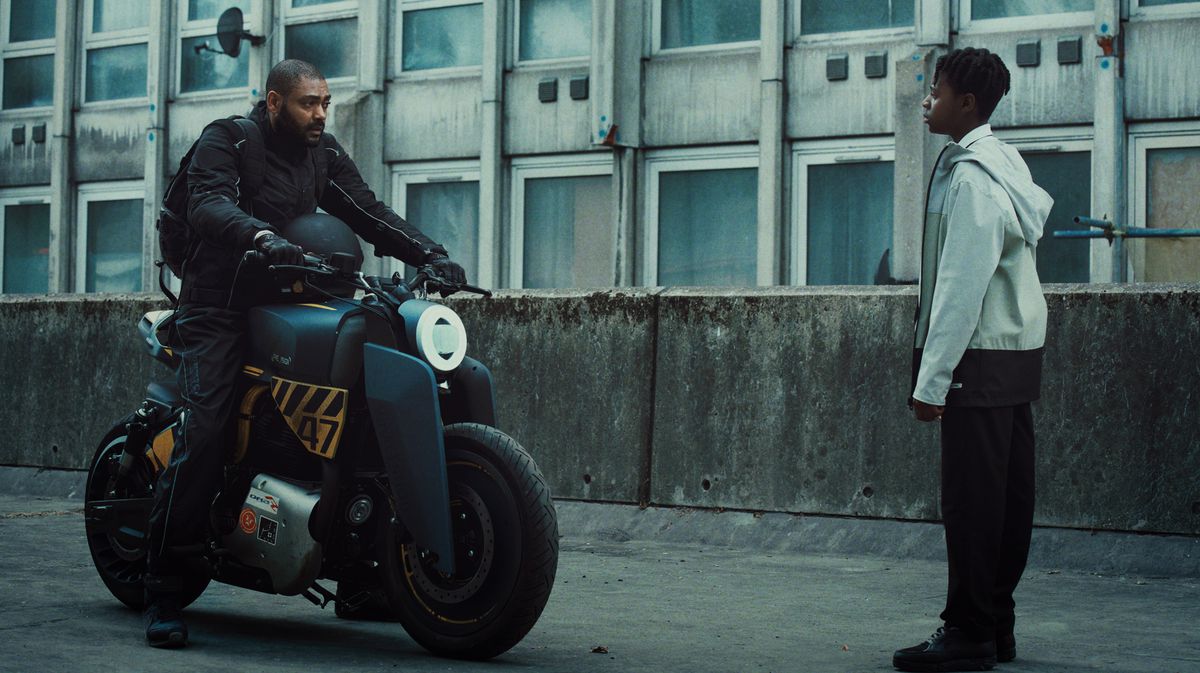 (LR) גבר בחליפת אימונית שחורה (קיין רובינסון) על גבי אופנוע עתידני עומד מול ילד צעיר עם קפוצ'ון לבן (ג'דייה באנרמן) ומכנסיים שחורים במטבח.