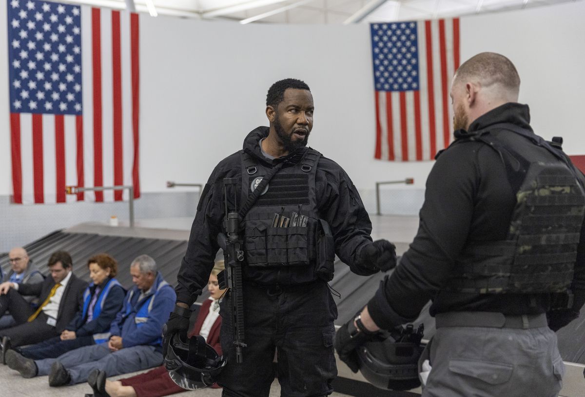 Майкл Джей Уайт в бронежилете и с винтовкой на плече разговаривает с другим мужчиной в бронежилете, в то время как заложники выстраиваются в очередь у багажной ленты в аэропорту в фильме «Еще один выстрел».