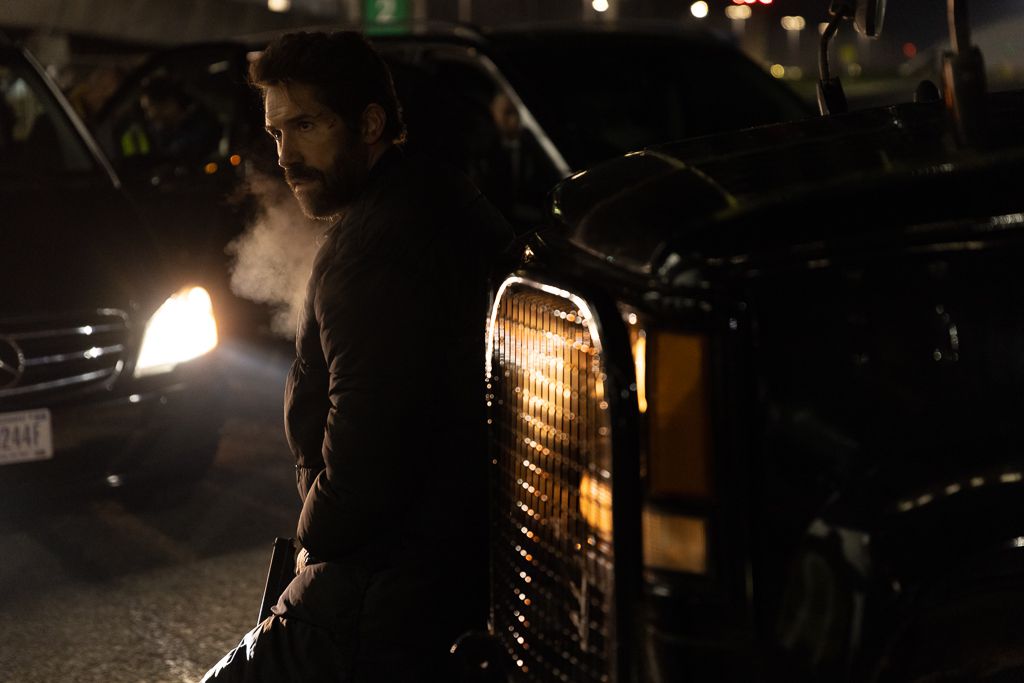 Скотт Адкинс прислоняется к передней части машины, и вы можете видеть, как он выдыхает в холодную погоду в фильме «Еще один кадр».