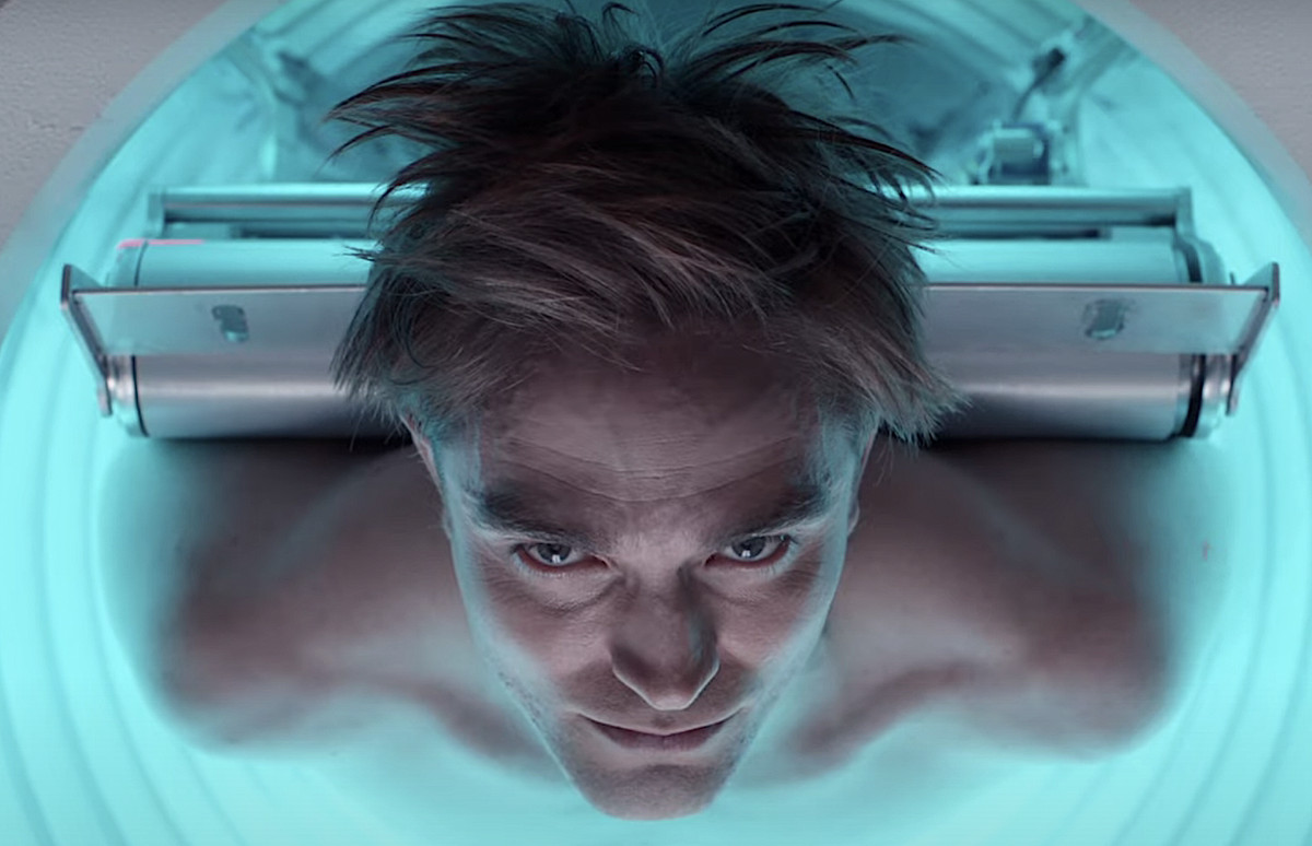 Robert Pattison ligt op zijn rug, zonder shirt, in een gloeiende blauwe MRI-achtige machine, rechtstreeks in de camera starend, in een vroege teaser-trailer voor Bong Joon-ho’s Mickey 17