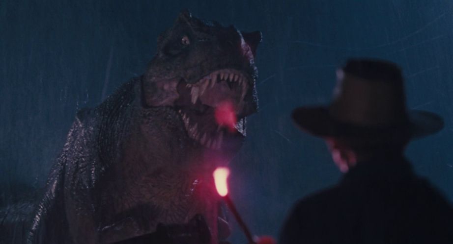 A man wearing a hat holds a flare in front of a roaring tyrannosaurus rex in Jurassic Park.