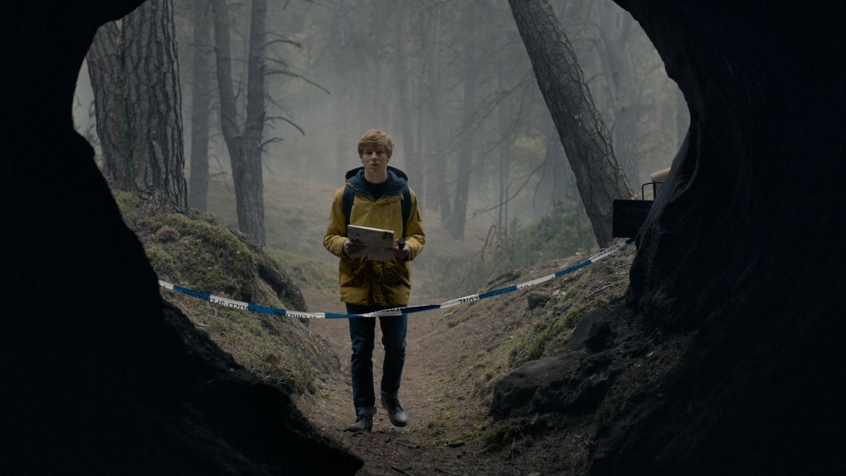 צעיר לבוש במעיל גשם צהוב עומד מול הכניסה המחולקת של מערה גדולה ביער בחושך.