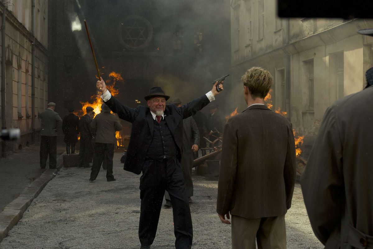 Ein Mann mit Hut und weißem Spitzbart ohne Schnurrbart hebt triumphierend die Arme vor einer brennenden Synagoge. Er hält eine Waffe
