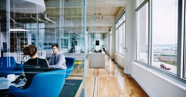 שני אנשים יושבים על כיסאות מדברים במשרד בקומה גבוהה עם חלונות גדולים מימין, נוף עירוני בנוף