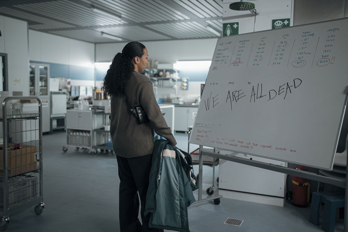 קאלי רייס בתור אוונג'לין נבארו עומדת מול לוח מחולק חלקית עם המילים "כולנו מתים", בבלש אמיתי: ארץ לילה.