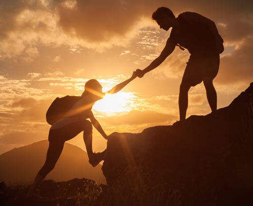 One person helps another ascend a mountain climb while the sun rises or sets in the background