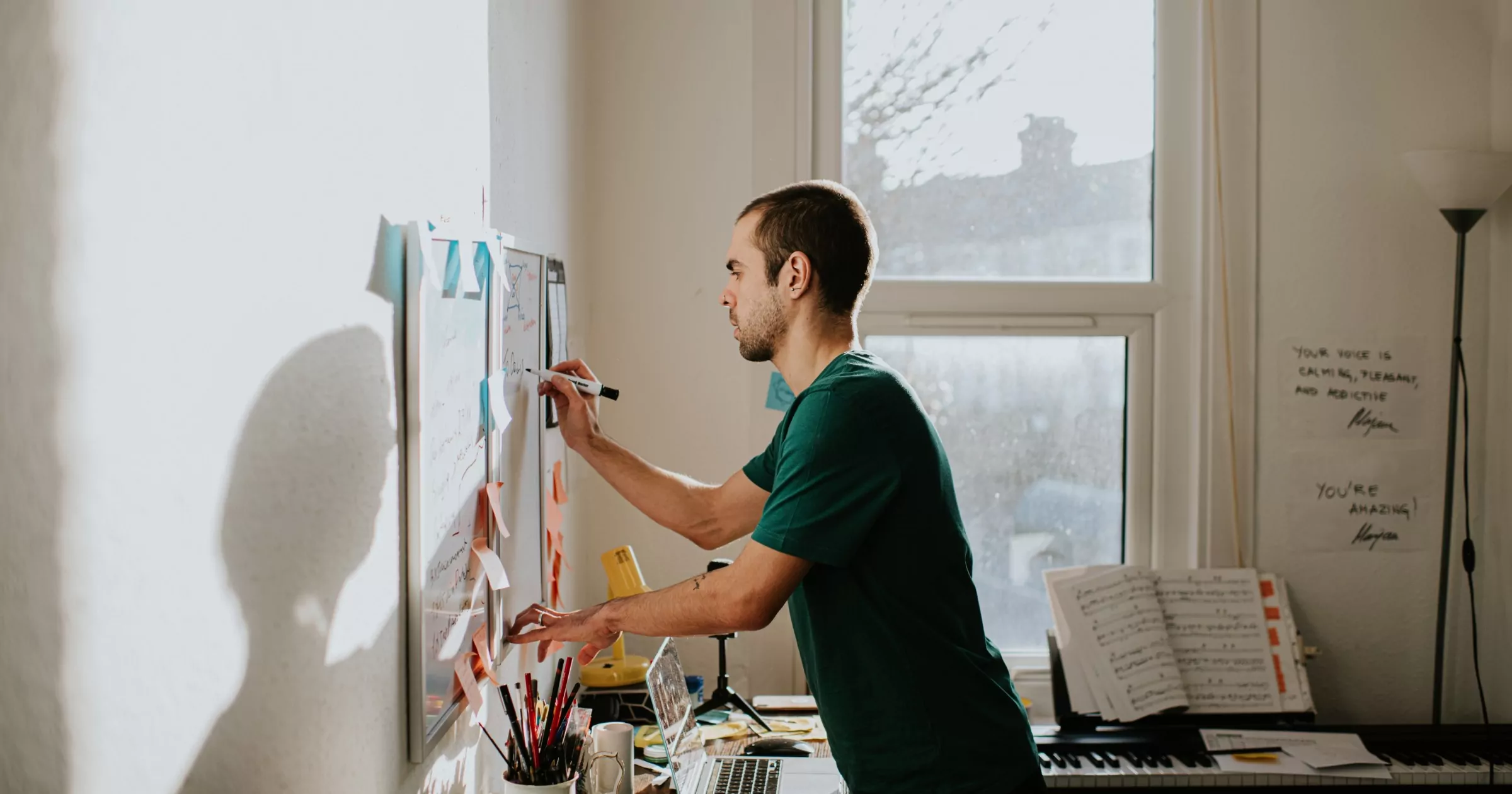 A man leans over a messy desk and writes on a wall-mounted whiteboard in a home-office environment