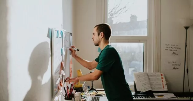 En mand læner sig over et rodet skrivebord og skriver på en vægmonteret tavle i et hjemmekontormiljø