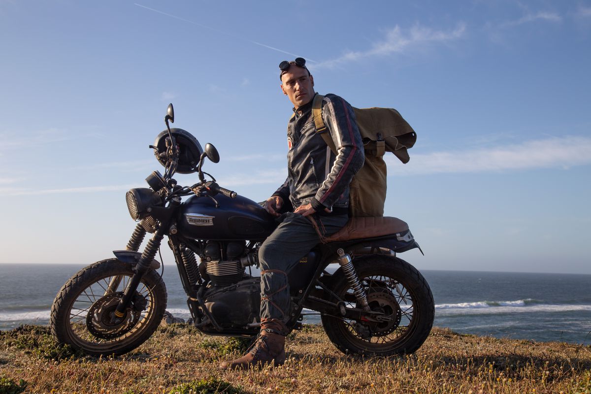 Marko Zaror fica descolado em uma motocicleta, vestindo uma jaqueta de couro e óculos de proteção no topo da cabeça, em Fist of the Condor, com o oceano atrás dele.